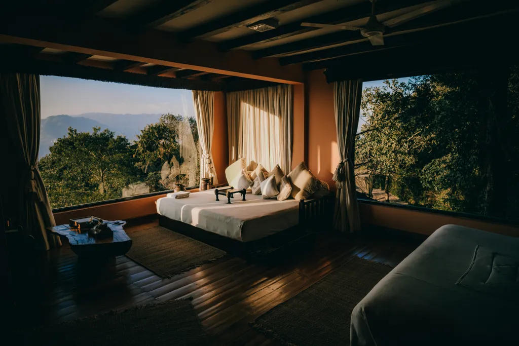 A hotel room at golden hour, its wide windows open onto forested hills and a distant ridge